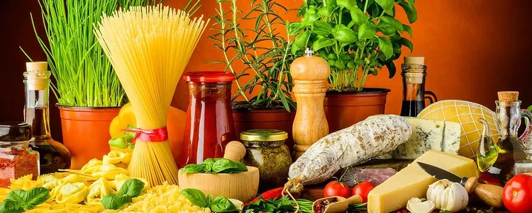 A photo of italian cooking ingredients. Tomatoes, pasta bundles, sausage, parmesan cheese wheels, pepper mills, the whole nine yards.