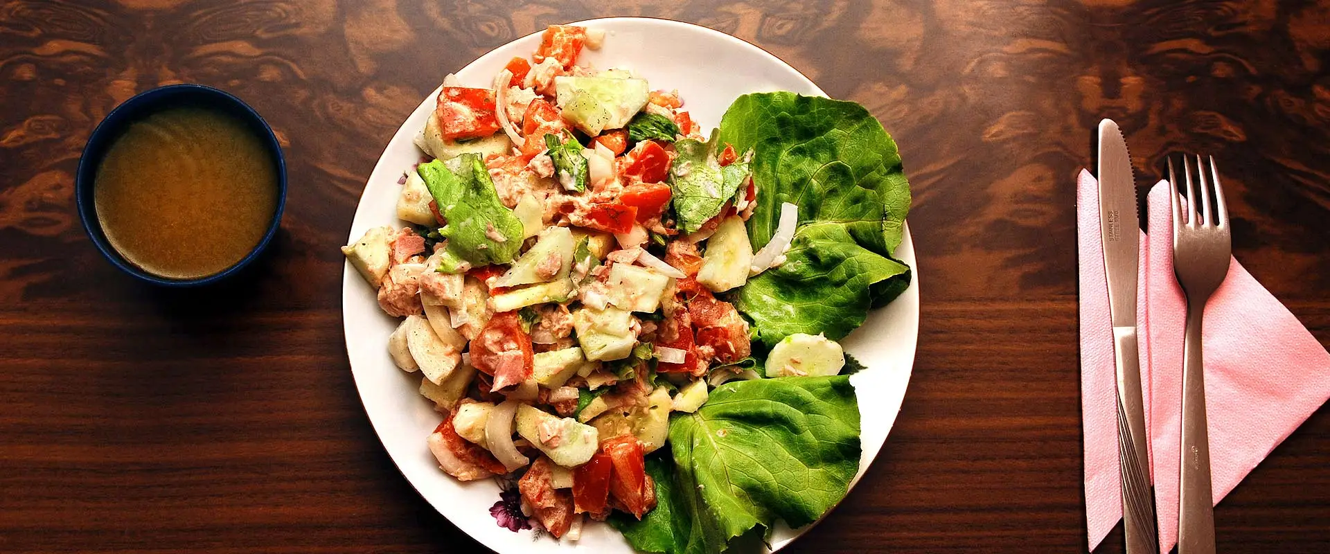 A photo of a delectable greek salad with two large lettuce leaves used as garnish. The plate is on a smooth wood table beside a small bowl of warm gravy on the left and utensils on the right