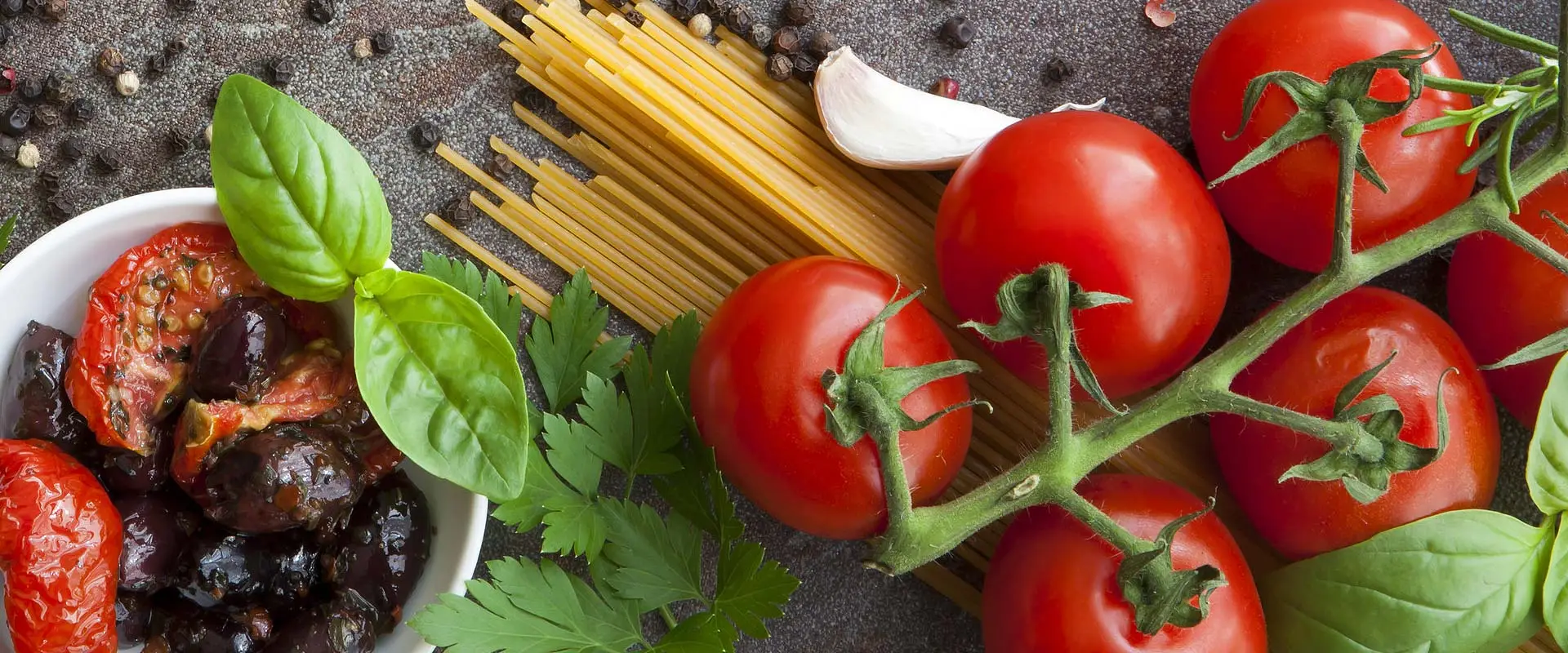 A photo of the raw ingredients for spaghetti. Firm ripe tomatoes, long rigid noodles, dried garlic and an aromatic sprig of oregano.